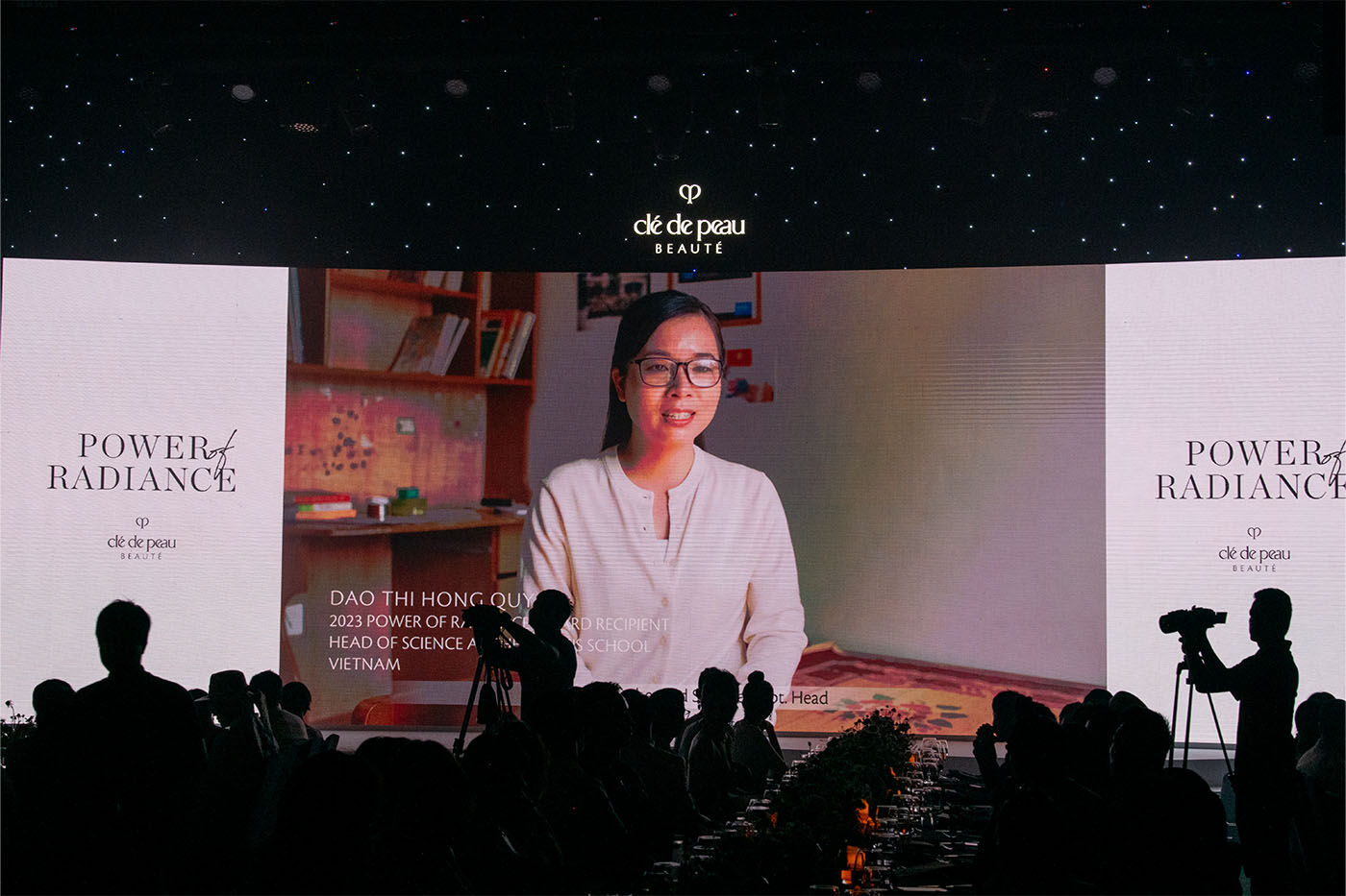 Photo of a video of Ms. Dao Thi Hong Quyen displayed on a large screen with silhouettes of attendees watching at the 5th anniversary of the Power of Radiance Awards.