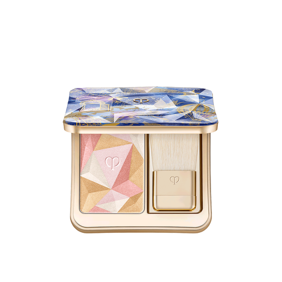Photo of the 26 AW Holiday Collection limited-edition THE LUMINIZING FACE ENHANCER. The opened gold compact reveals a multi-colored geometric powder and a brush, under a blue and purple prismatic lid with gold illustrations.
