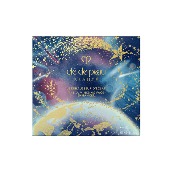 Photo of the 26 AW Holiday Collection limited-edition THE LUMINIZING FACE ENHANCER outer box. The square package features a scenic illustration of Earth from space under a starry night sky, with a large gold shooting star across the top.