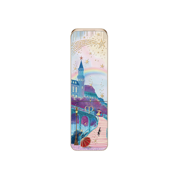 Photo of the 26 AW Holiday Collection limited-edition LIPSTICK case. The closed rectangular gold tube features a scenic illustration of a cobblestone path toward a castle with a rainbow, detailed with figures, a black dog, and a red umbrella.
