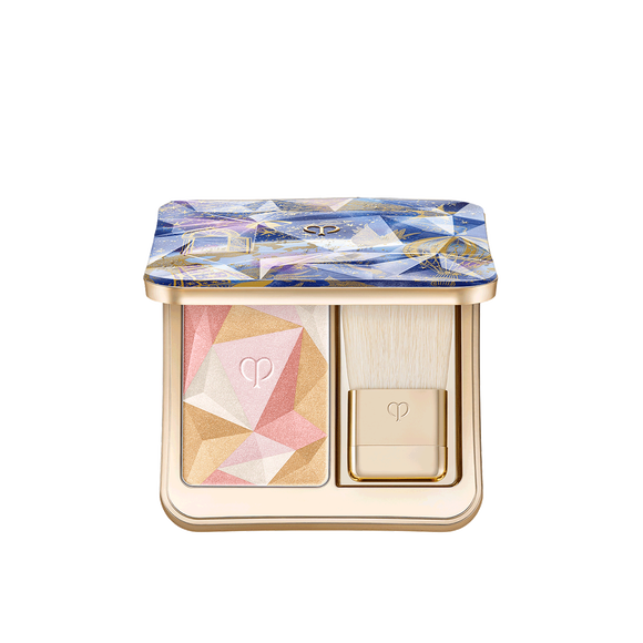 Photo of the 26 AW Holiday Collection limited-edition THE LUMINIZING FACE ENHANCER. The opened gold compact reveals a multi-colored geometric powder and a brush, under a blue and purple prismatic lid with gold illustrations.