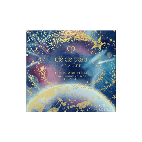 Photo of the 26 AW Holiday Collection limited-edition THE LUMINIZING FACE ENHANCER outer box. The square package features a scenic illustration of Earth from space under a starry night sky, with a large gold shooting star across the top.