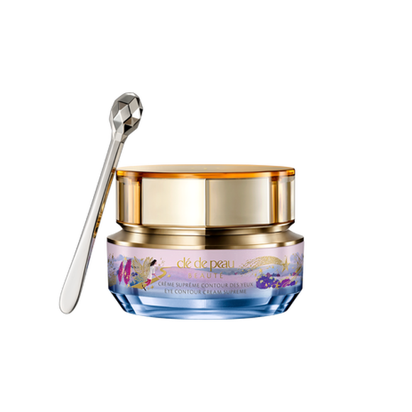 Photo of the 26 AW Holiday Collection limited-edition EYE CONTOUR CREAM SUPREME. The gold-trimmed jar features a blue and pink pastel illustration with a gold shooting star, shown beside a faceted silver spatula.