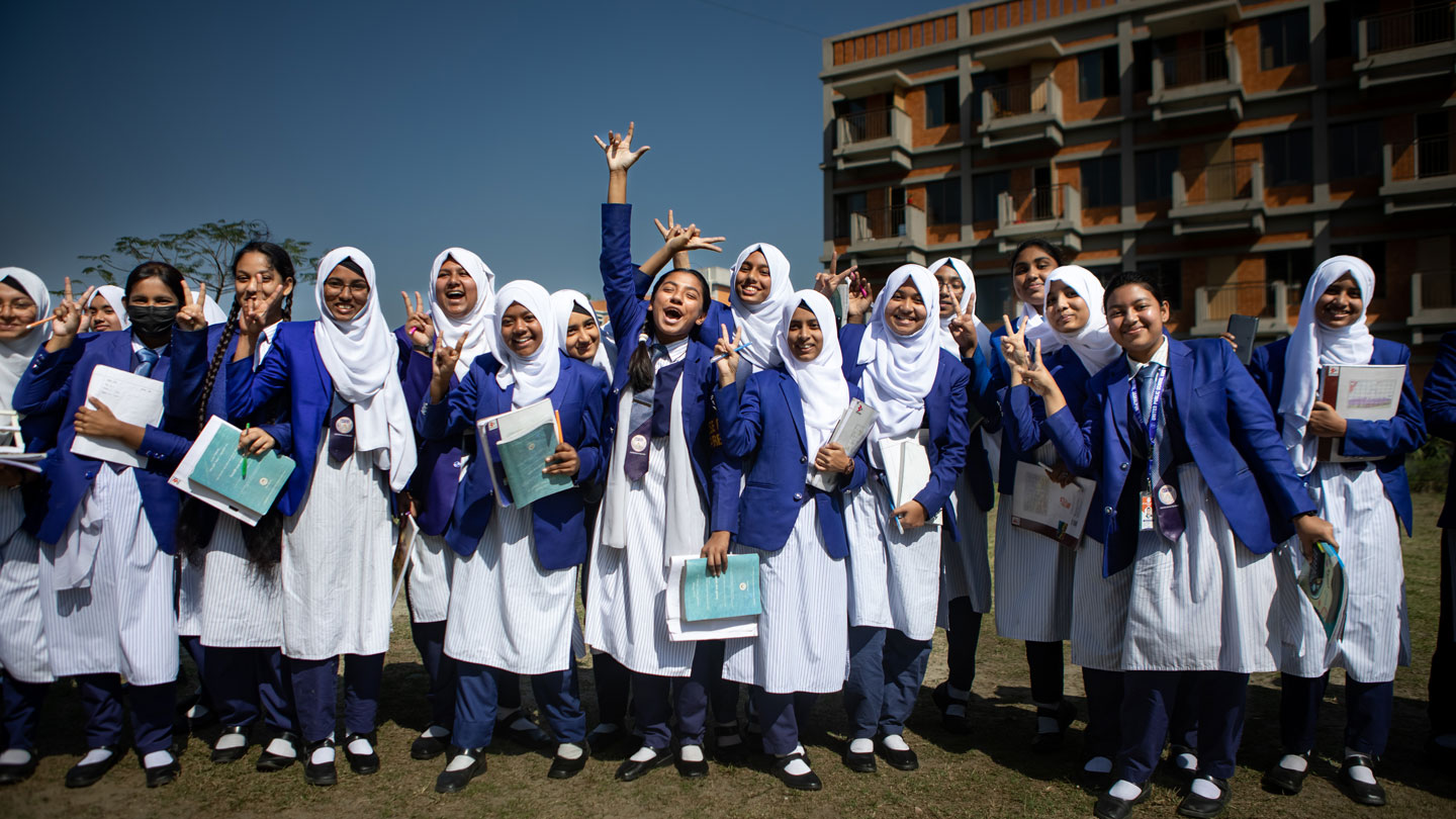 Photo of Bangladeshi schoolgirls in uniform smiling and holding textbooks outside their school building.