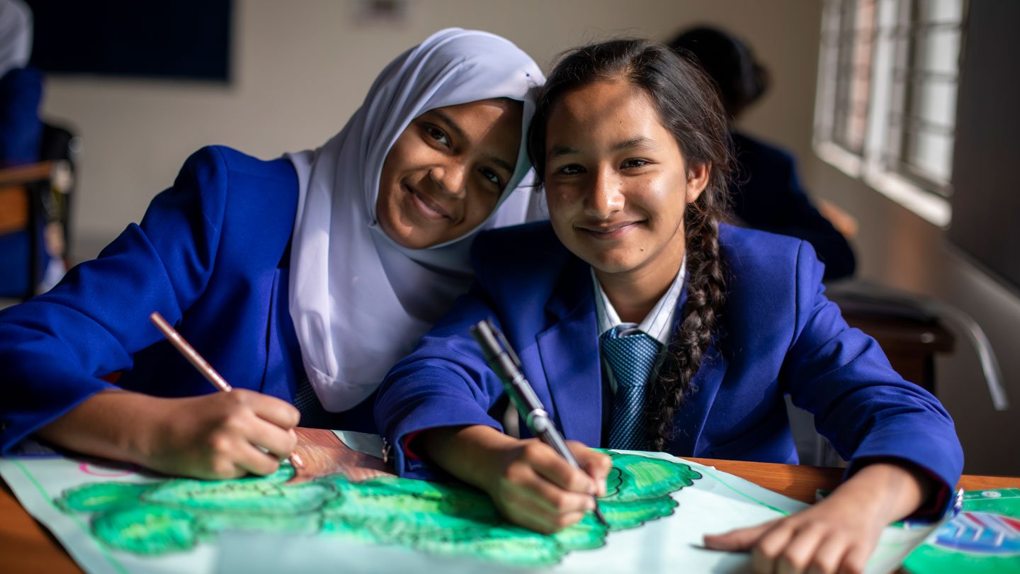 Photo of two girls smiling at the camera while painting as part of a classroom activity in the new curriculum.
