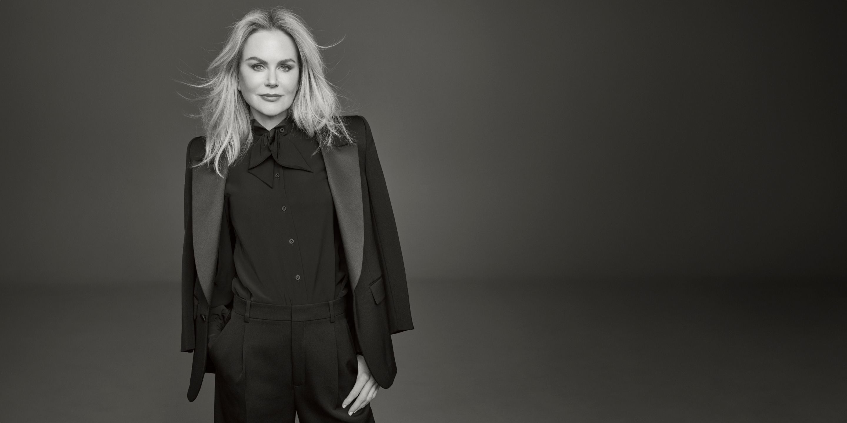 Photo of Clé de Peau Beauté’s new global brand ambassador, Nicole Kidman, wearing a black suit with right hand in pocket, looking directly at the camera in a monochrome image.