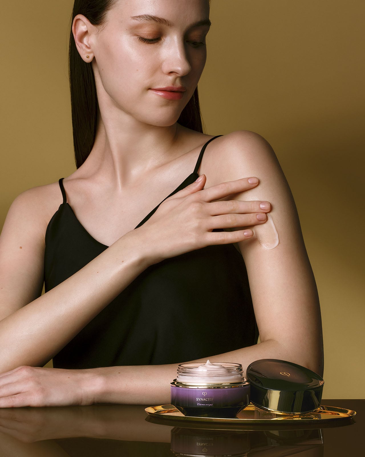 Photo of a light skin tone model applying SYNACTIF body cream to her arm. An open jar of the cream sits on a gold tray in the foreground against a gold background.