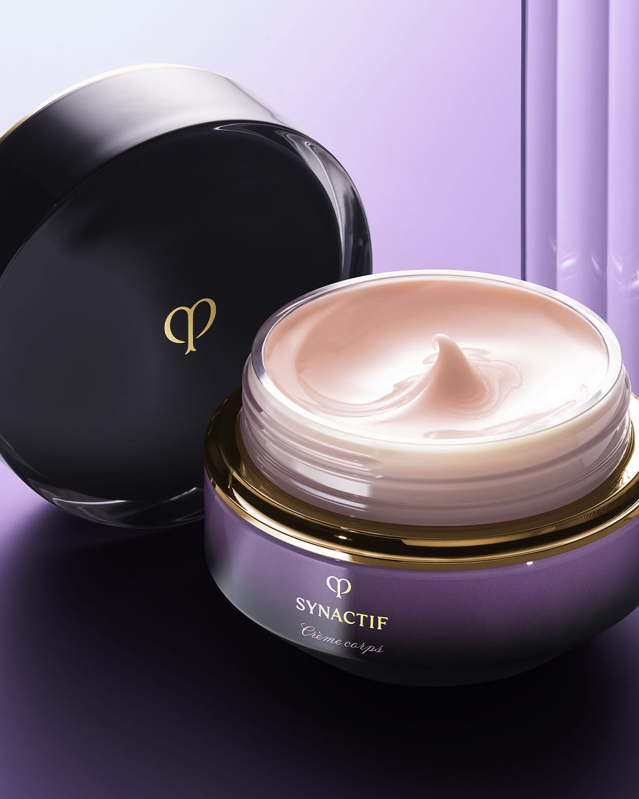 Photo of an open jar of SYNACTIF body cream, showing the soft, pale pink cream inside with its black cap leaning against it. The scene is set against a soft purple and white gradient background.